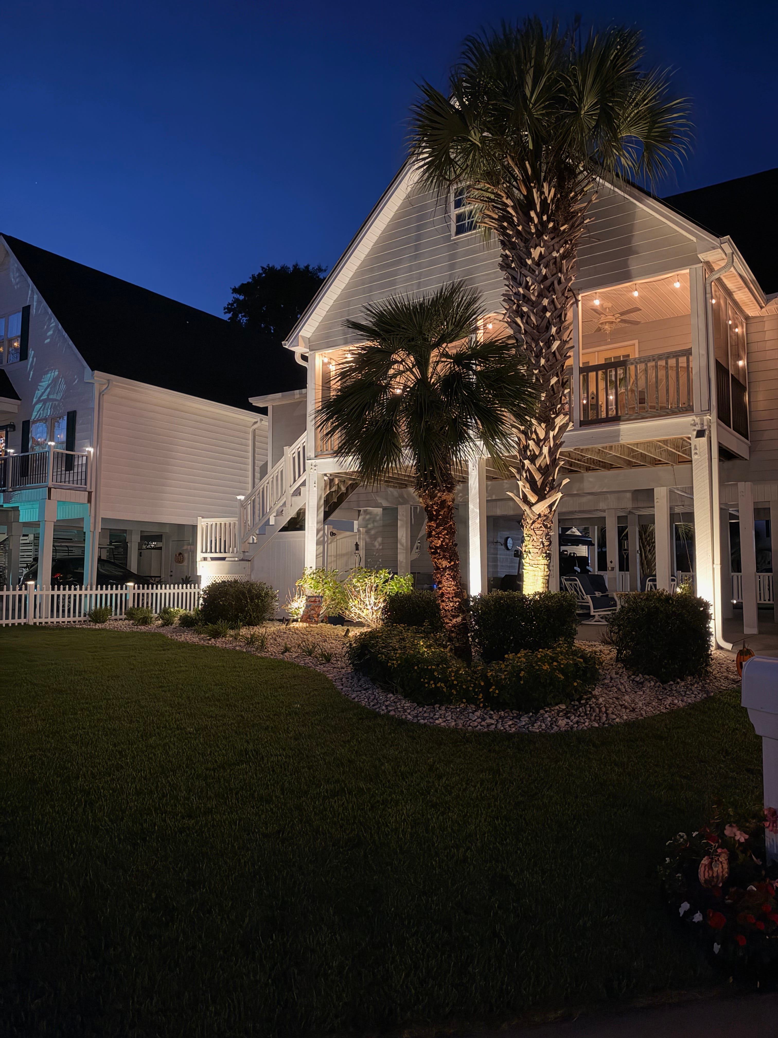 Nighttime landscape lighting on palms, garden beds, and a coastal home